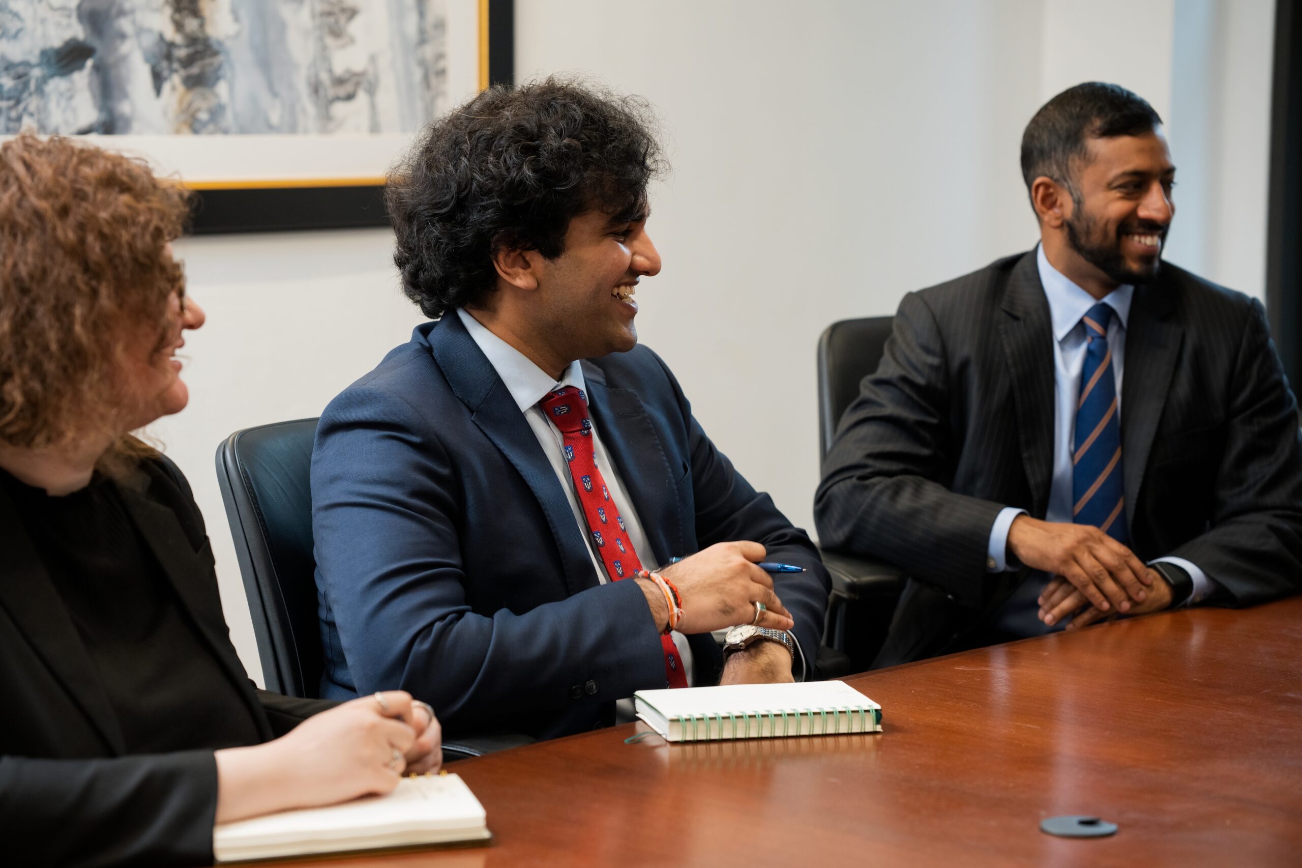 Three professionals in business attire sit around a conference table, smiling and engaged in a discussion, with notebooks and a pen visible in a modern office setting